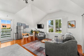 A spacious living area with a grey sofa, wooden dining table, and chairs, a flat-screen TV mounted on the wall, and a large window offering a view of the outdoors. - Carriage House at Historic Chapman House Arcata (Arcata)