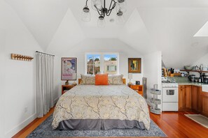 Une chambre avec un grand lit, une fenêtre, un coin cuisine et un lustre.