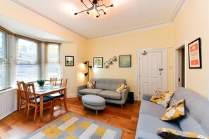 Ein Wohnzimmer mit einer Couch, einem Hocker und einem Esstisch mit Stühlen. An der Wand hängen gerahmte Bilder und ein Türgriff ist zu sehen.