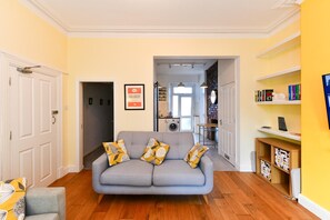 Ein Wohnzimmer mit grauem Sofa, einem Bücherregal und einer durch einen offenen Durchgang sichtbaren Küche.