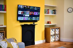 A living room with a flat-screen TV mounted on a yellow wall, a fireplace, shelves with books, and a clock on the wall. - Charming Apartment in the Heart of Llandaff - Pass the Keys (Cardiff)