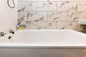 Et badeværelse med et hvidt badekar, brusehoved og en shampoo-flaske.