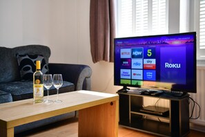 A living room with a television displaying the Roku interface, a bottle of wine, and two glasses on a wooden table. - Spacious Cardiff Apartment | WiFi | Near City - Pass the Keys (Cardiff)