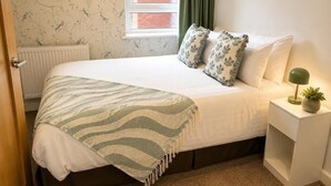 Una cama bien hecha con una colcha a rayas, dos almohadas con estampado floral, una lámpara de noche y una pequeña planta en maceta sobre una mesita de noche blanca.