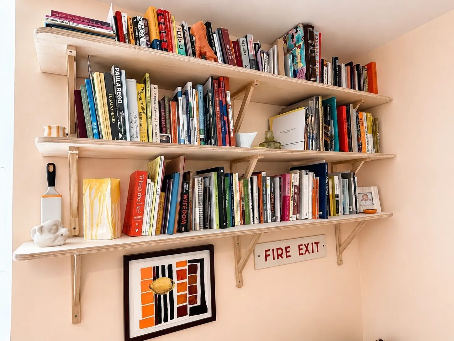 Une bibliothèque à plusieurs étagères remplie de livres, une œuvre d’art encadrée et un panneau indiquant la sortie de secours.