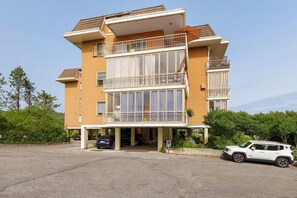 Un immeuble résidentiel à plusieurs étages, doté de balcons, d’un espace de stationnement couvert et d’un VUS blanc garé devant.