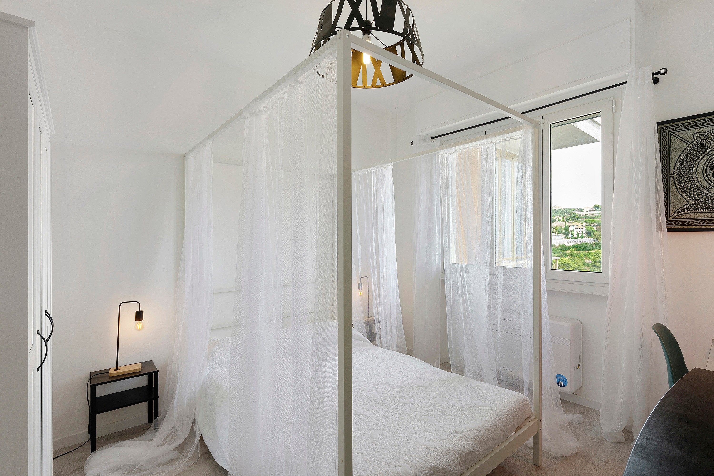 Ein Schlafzimmer mit Baldachinbett, Nachttisch mit Lampe und Fenster mit Blick auf Grünflächen.