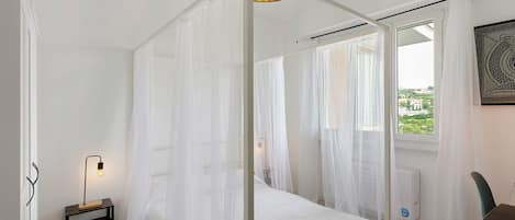 A bedroom with a canopy bed, a bedside table with a lamp, and a window with a view of greenery.