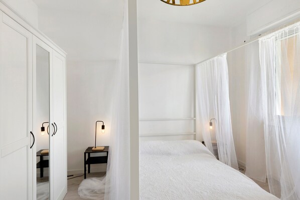 A bedroom with a large bed, a bedside table, a mirror, and sheer curtains.