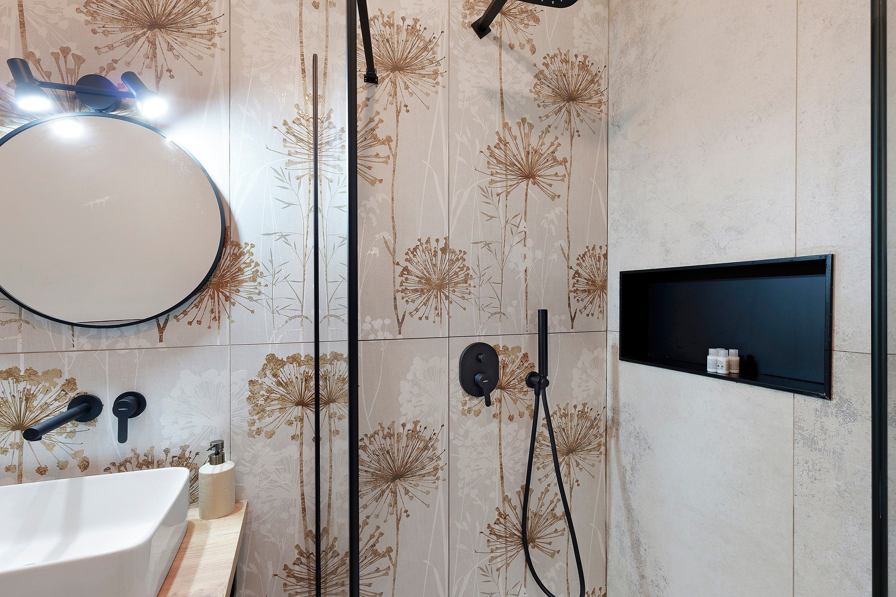 Une salle de bain avec un miroir rond, un lavabo, une douchette à main et une étagère avec des bouteilles.