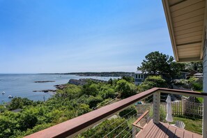 A coastal view from a balcony with a wooden deck, overlooking a bay with boats and a distant shoreline. - Vista Mare: Hilltop Home with Ocean Views (Rockport)