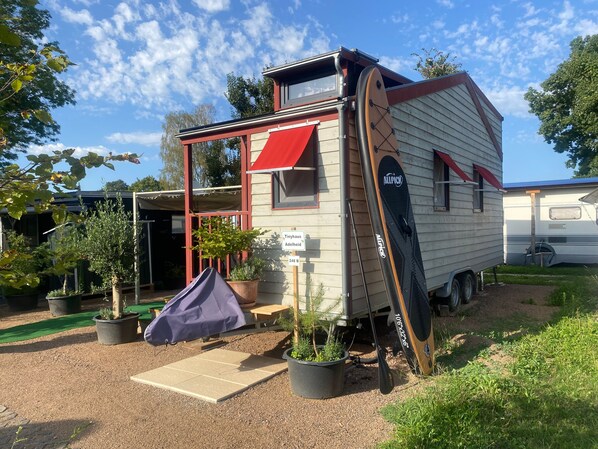 Una casa móvil con porche cubierto, puerta corrediza y un pequeño jardín con plantas en macetas.
