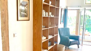 A room with a wooden bookshelf, a framed picture on the wall, a blue chair, and a window with a view of greenery.