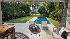 Un espace aménagé au bord de la piscine, offrant une vue sur celle-ci, entouré d’une végétation luxuriante et doté de fauteuils en osier et d’une table en bois.