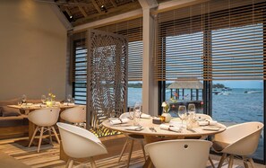 A dining area with a view of the sea, featuring a round table set with white chairs, a decorative partition, and a thatched-roof structure visible through the windows. - "VILLA PLENITUDE ANAHITA" | Private pool, golf course view, access to the resort (Beau Champ)
