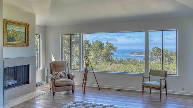 A spacious room with a large window overlooking a coastal landscape, a fireplace, and a comfortable seating area. - Pebble Beach Escape - Golf, Ocean & Sunset Views (Pebble Beach)
