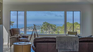 A room with a large window overlooking a coastal landscape, a leather sofa, a chair, and a tripod.