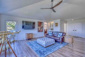 A spacious living room with a ceiling fan, a mounted television, a leather sofa, a dining area with a glass table, and a patterned rug.