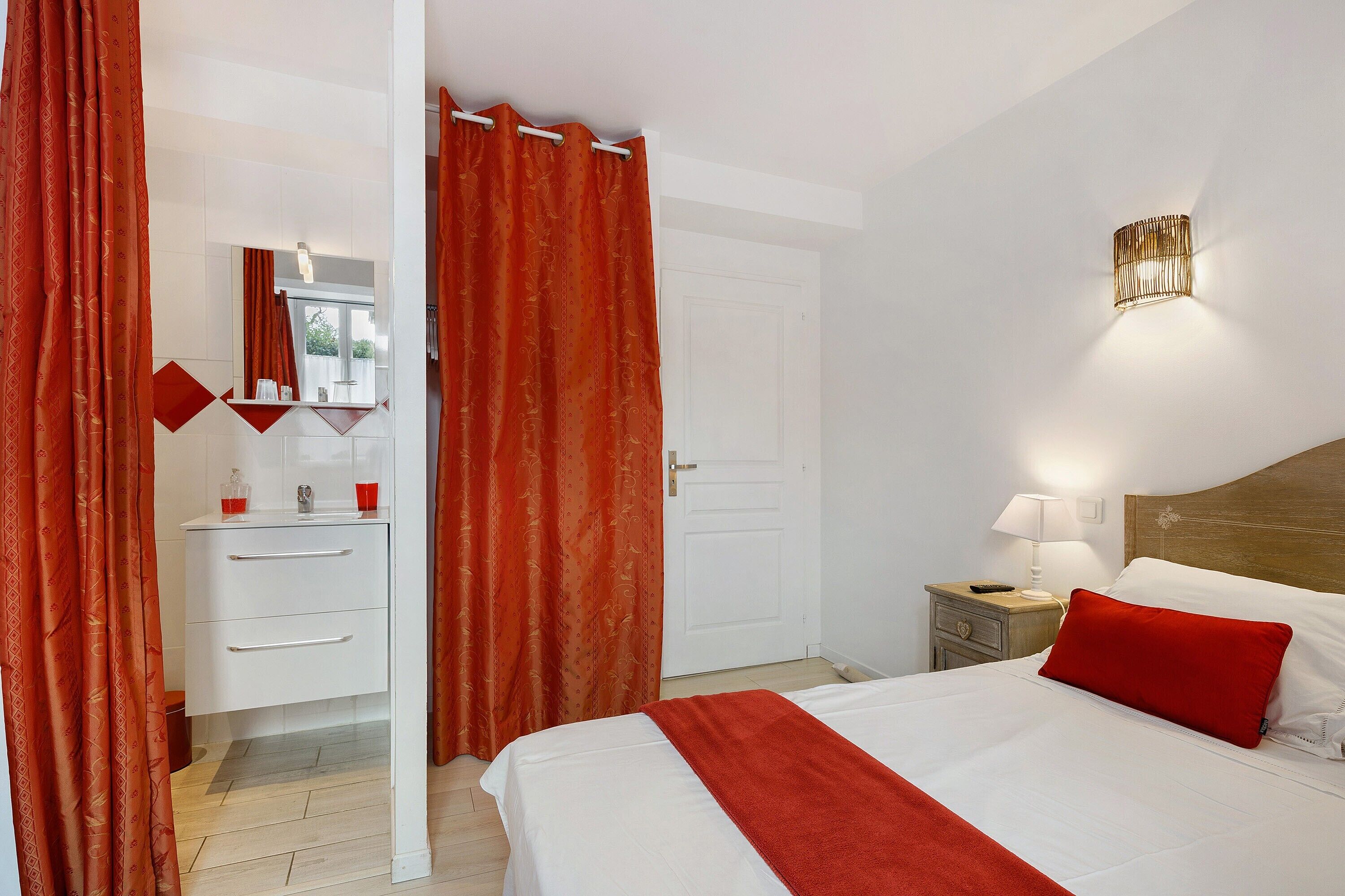 Une chambre d’hôtel avec un lit, une table de chevet, une lampe, un miroir, un lavabo et des rideaux rouges.