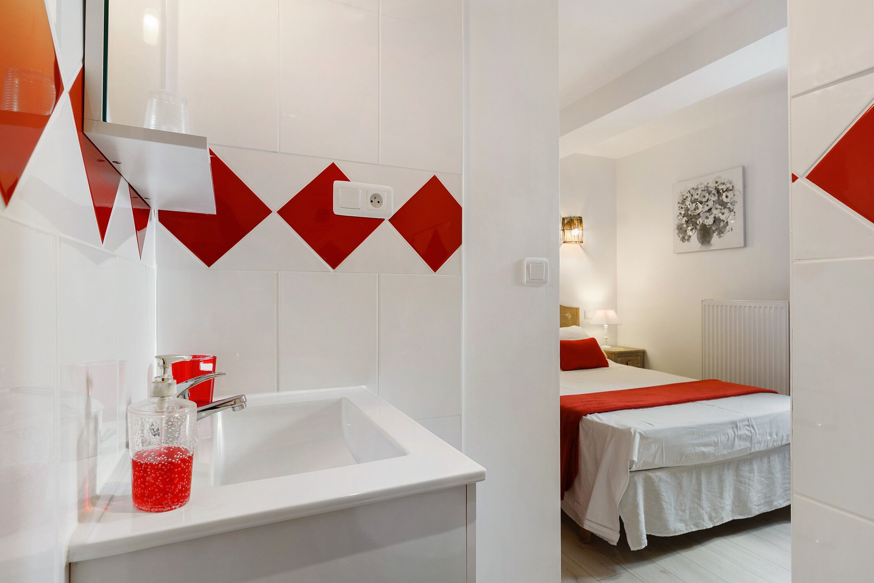 A hotel room with a white sink, red accents, a bed with white bedding, a nightstand, and a wall-mounted light fixture.