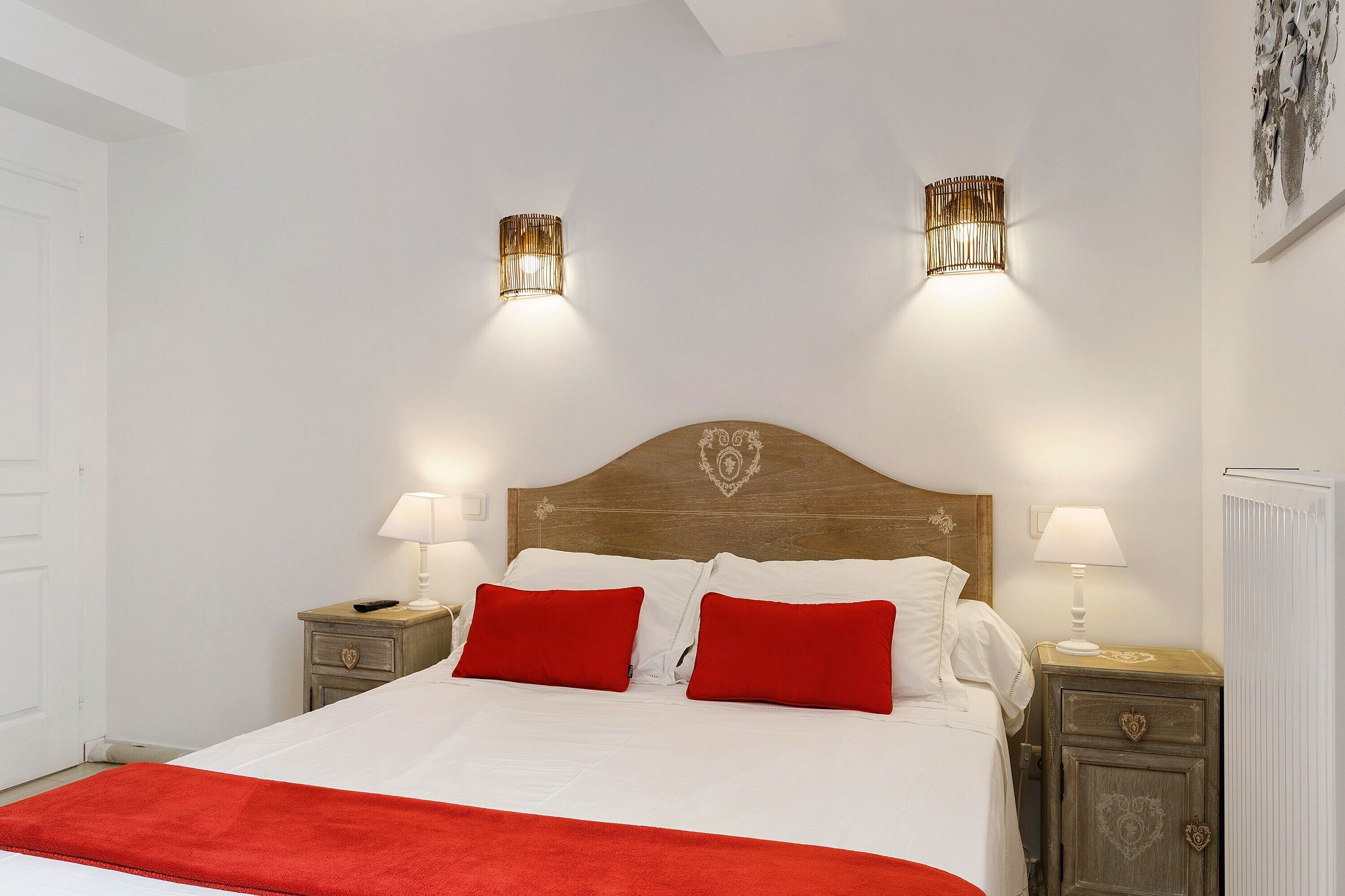 Un dormitorio con cabecera de madera, dos lámparas de noche, cama con ropa de cama blanca y almohadas rojas, y una manta roja.