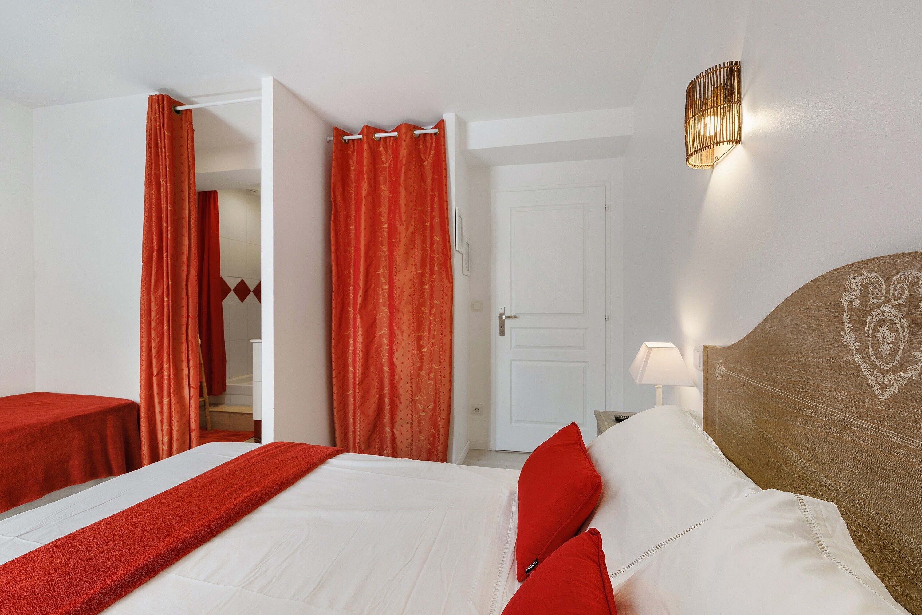 A hotel room with a bed, a nightstand, a lamp, a door, and red curtains.