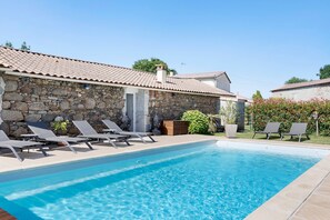 Un edificio de piedra con techo de tejas, piscina y sillones de descanso.