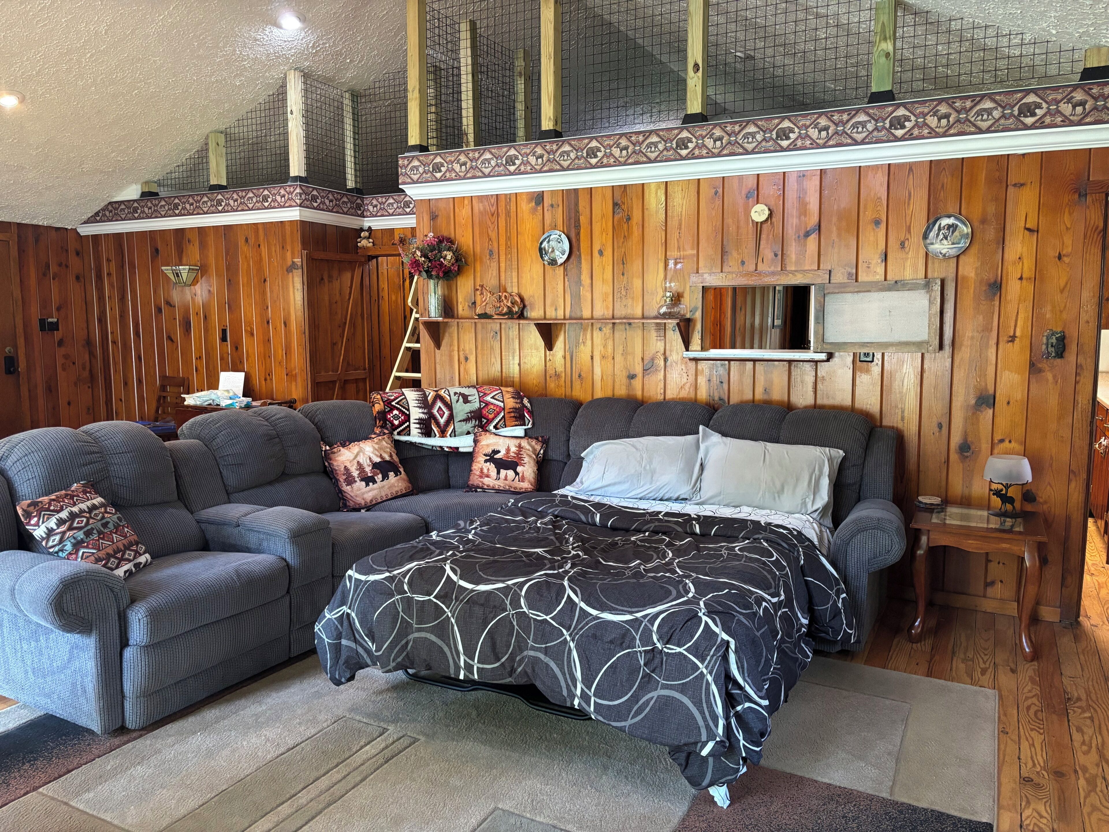 Ein gemütliches Zimmer mit einer Holzwand, einer Couch, einem Bett mit gemusterten Betttextilien und einem kleinen Tisch mit einer Lampe.