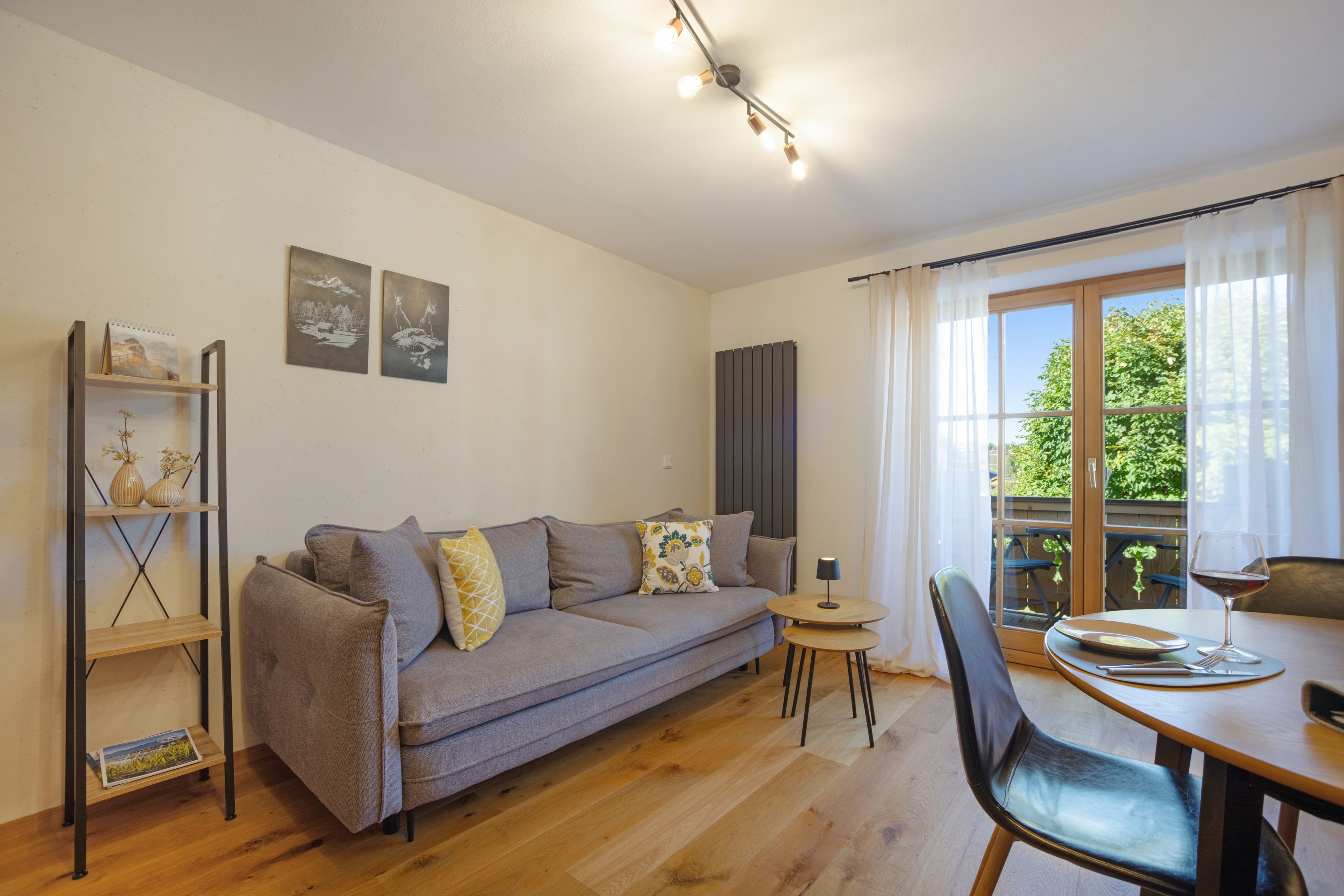 Een moderne woonkamer met een grijze bank, een houten salontafel en een eettafel met stoelen. Twee wanddecoraties en een radiator zijn aanwezig.