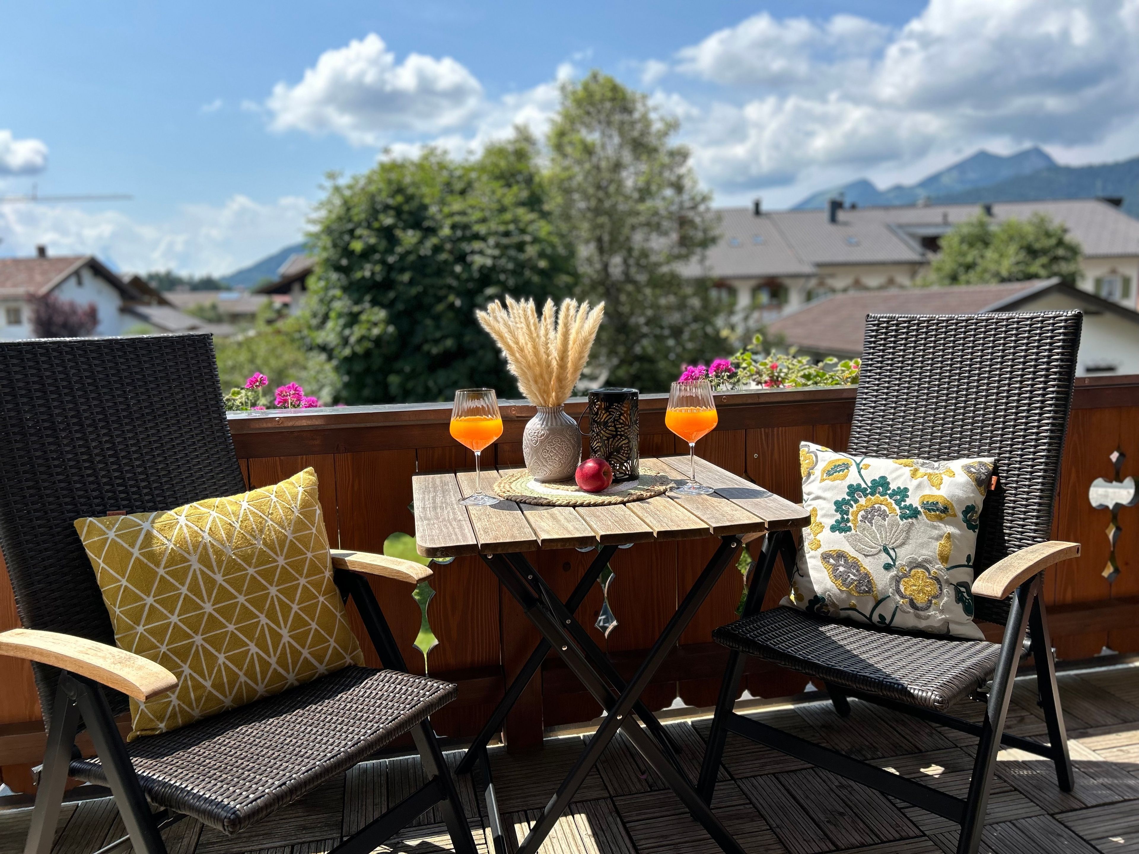 Eine Holzterrasse mit zwei Korbstühlen und einem Tisch, eine Vase mit Pampasgras, zwei Getränke und Blick auf Häuser und Berge.