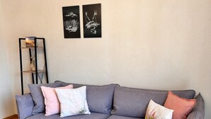 Un divano grigio con molti cuscini, un piccolo tavolino rotondo in legno con una pianta e una libreria nera addossata a una parete spoglia.