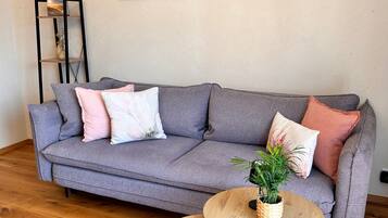Ein graues Sofa mit vielen Kissen, ein kleiner runder Holztisch mit einer Pflanze und ein schwarzes Bücherregal vor einer schlichten Wand.