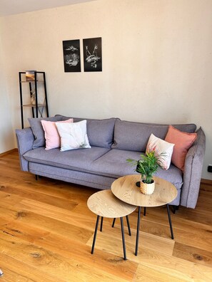 Ein graues Sofa mit vielen Kissen, ein kleiner runder Holztisch mit einer Pflanze und ein schwarzes Bücherregal vor einer schlichten Wand.