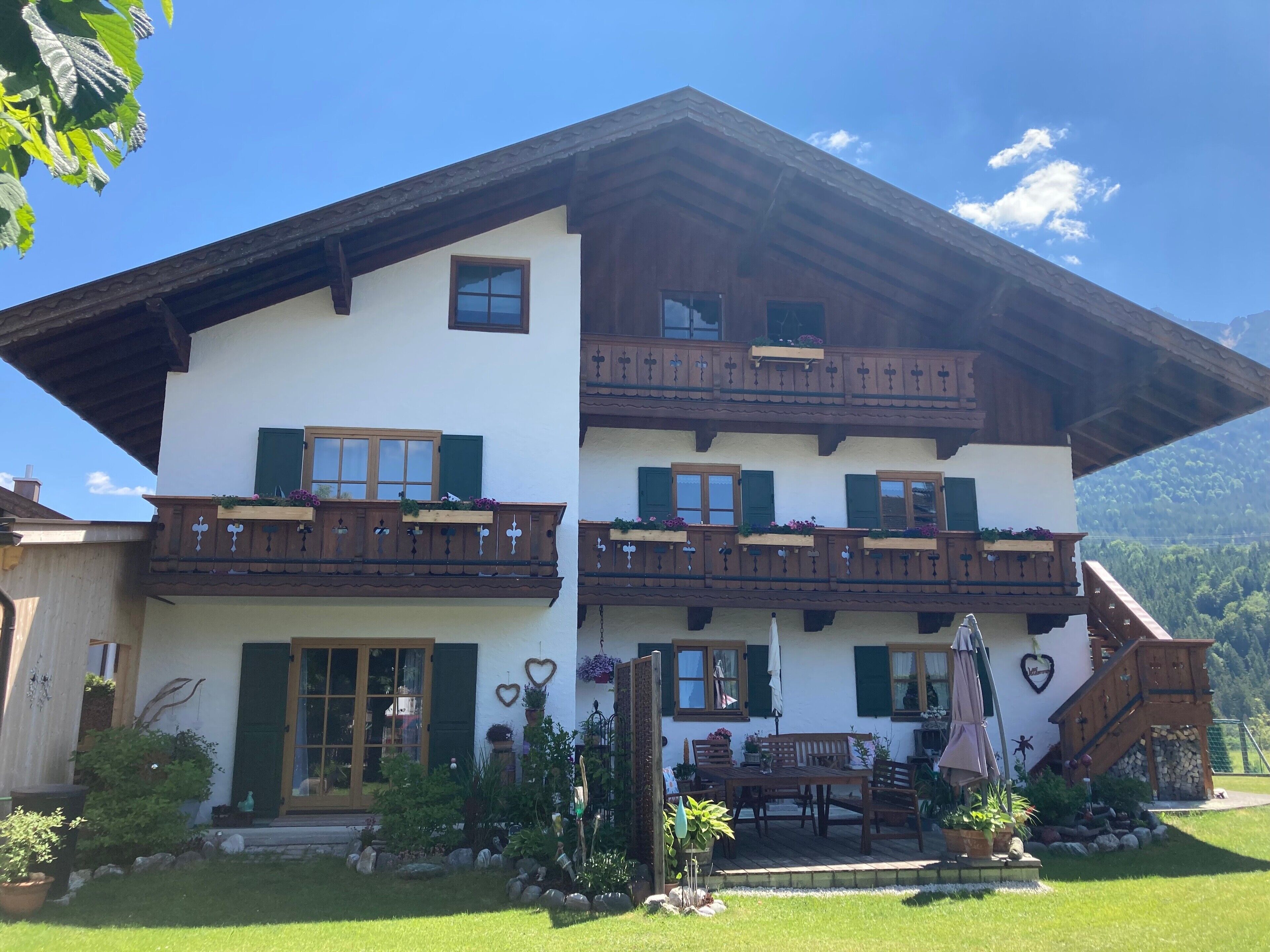 A two-story chalet with wooden balconies, a front entrance, and a garden area with outdoor furniture.
