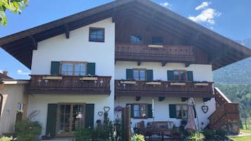 A two-story chalet with wooden balconies, a front entrance, and a garden area with outdoor furniture.