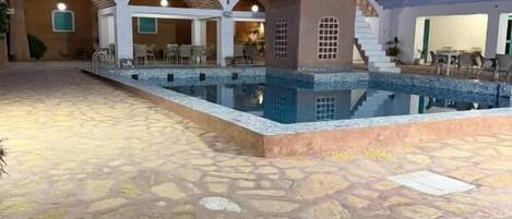 A pool area with a stone floor and a building with arches and a staircase.