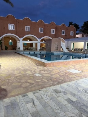 A pool area with a stone floor and a building with arches and a staircase.