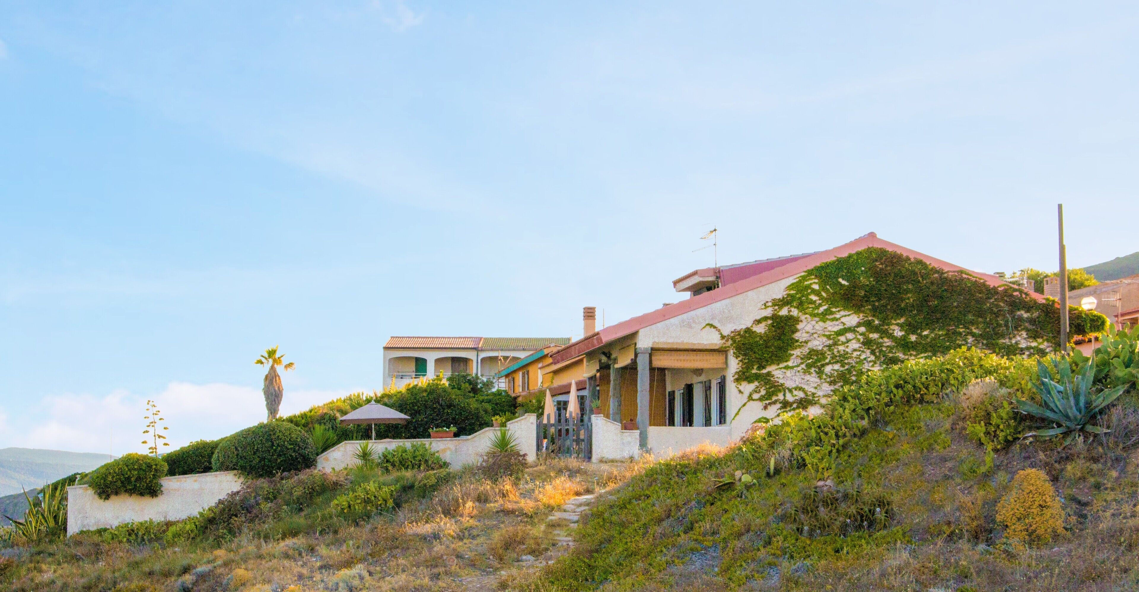 Une propriété située sur une colline, avec une maison dotée d’un toit rouge, d’un patio couvert et d’un jardin où poussent différentes espèces de plantes.