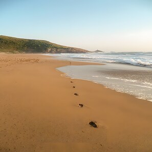 A sandy beach with footprints leading to the water, a grassy hill in the background, and waves approaching the shore. - Sardinia - House with Direct Beach Access, Sea View, Wi-Fi and Air Conditioning (Porto Alabe)