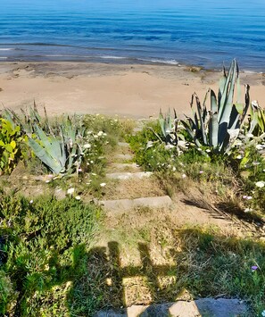 Ein Küstenpfad führt zu einem Sandstrand, begleitet von Agavenpflanzen und freiem Blick auf das Meer.