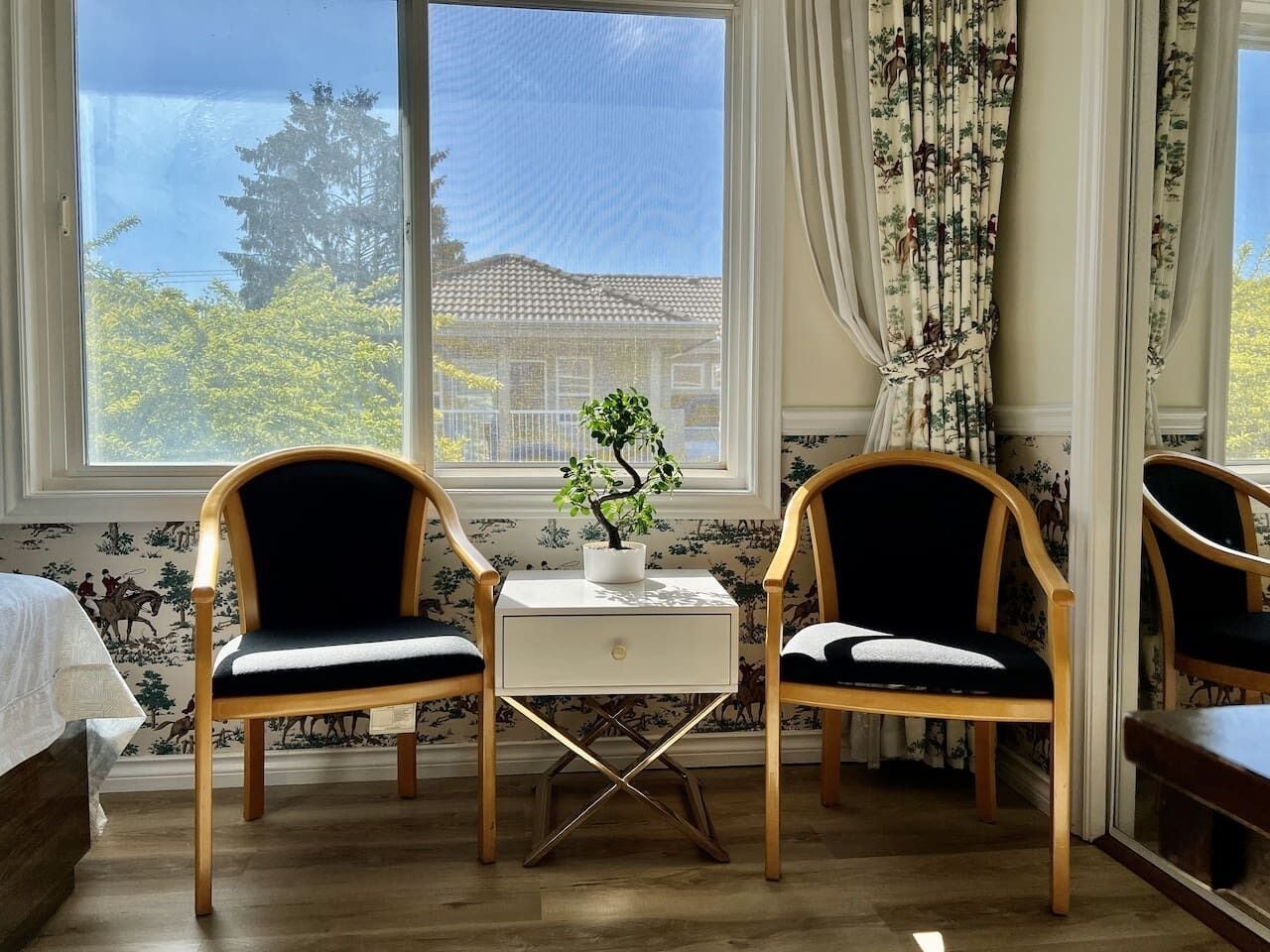 Een kamer met twee houten stoelen met zwarte kussens, een witte eettafel met een plant en een raam voorzien van gordijnen.