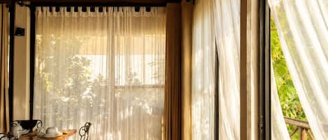 Una habitación con piso de madera, una mesa de comedor con sillas, una ventana con cortinas traslúcidas y vistas al exterior.