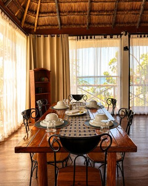 A dining area with a wooden table set for six, surrounded by chairs, with a bookshelf and curtains in the background.