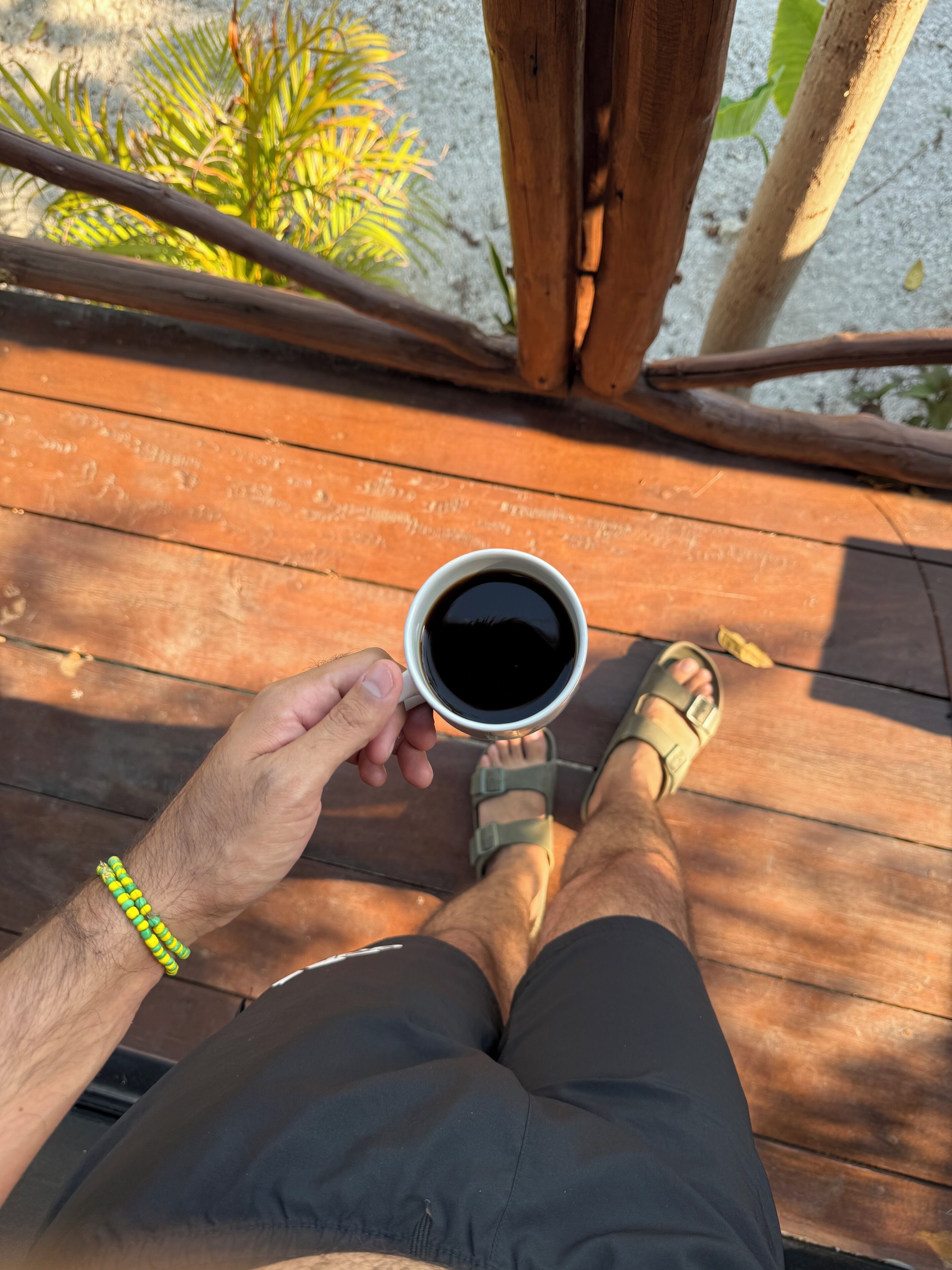 A person sitting on a wooden deck holding a cup of coffee, wearing sandals and shorts.
