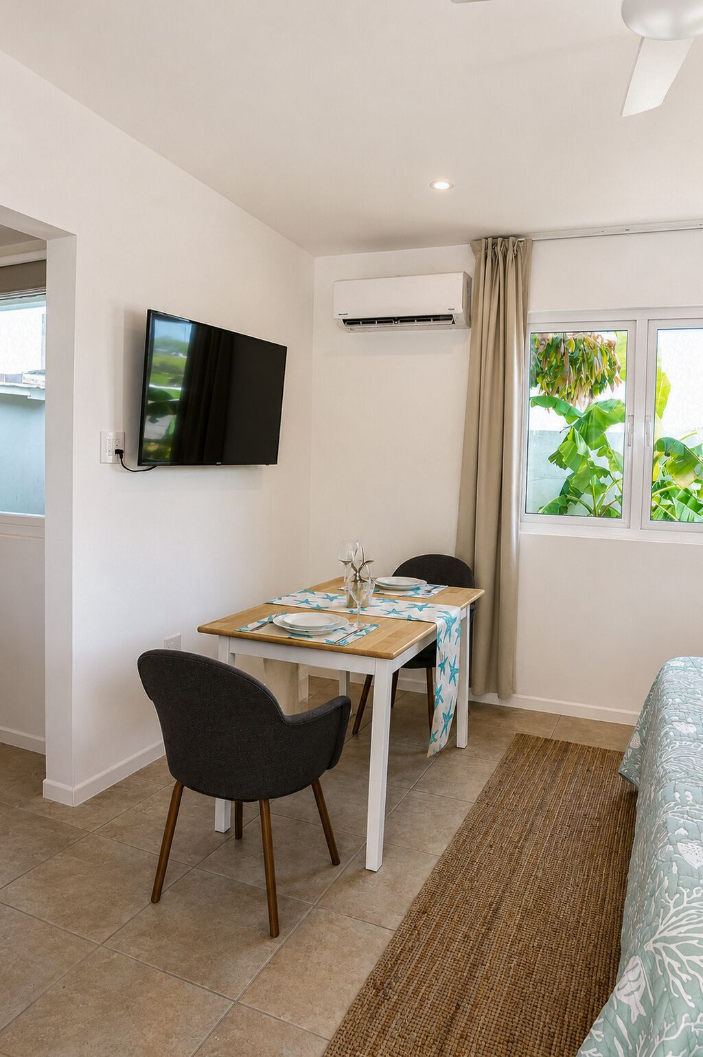 A room with a bed, a dining table with chairs, a TV mounted on the wall, and an air conditioning unit.