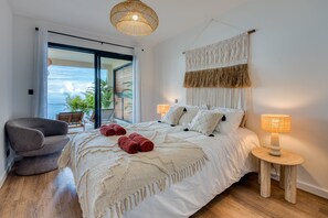Une chambre à coucher avec un lit, deux lampes de chevet, une chaise, une décoration murale en macramé et une vue sur l’extérieur.