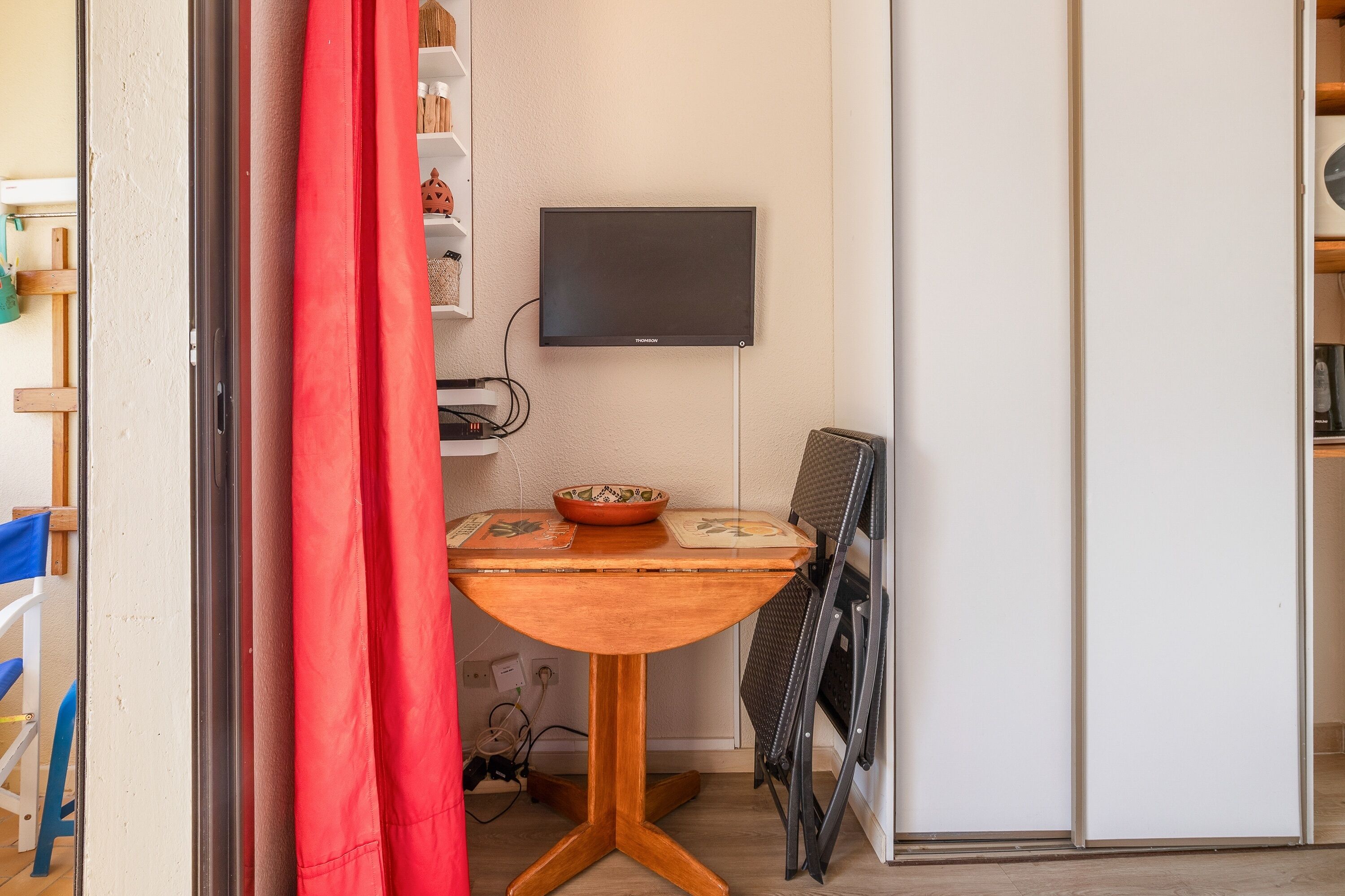 A compact room with a small table, a television mounted on the wall, and a red curtain partially drawn.