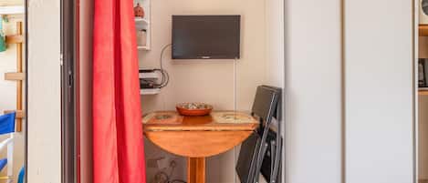 Een kleine kamer met een tafeltje, een aan de muur bevestigde televisie en een gedeeltelijk dichtgetrokken rood gordijn.