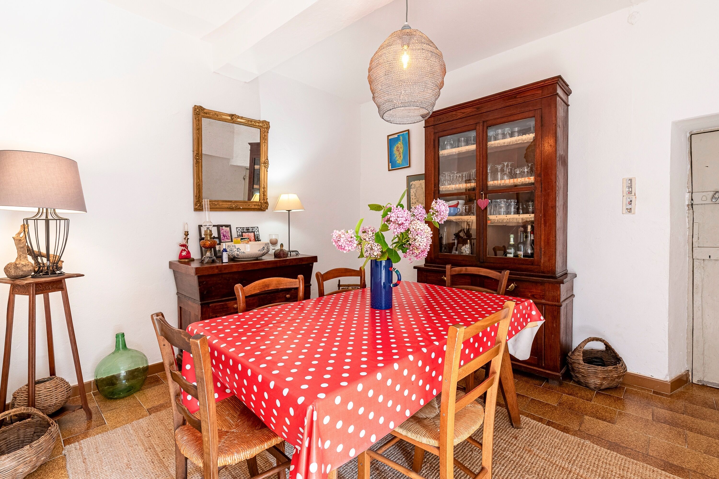 Un'area pranzo con tovaglia a pois rossi, sedie di legno, un vaso con fiori, un mobile vetrina e un lampadario.