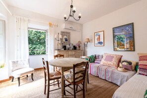Ein gemütliches Zimmer mit Esstisch, Stühlen, einer Couch und einem Bett. Es gibt ein Fenster mit Vorhängen, eine Chandelüster und Wandbilder.